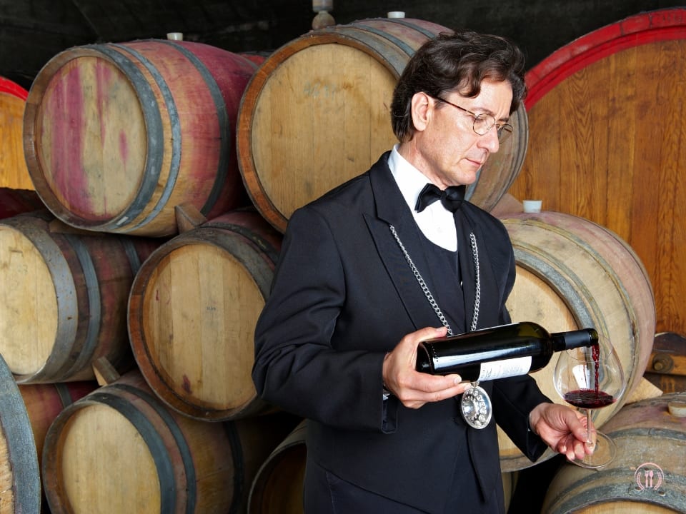 Head sommelier Filip Laskowski prezentuje kieliszek wina przy półce z butelkami w eleganckim wnętrzu restauracji.