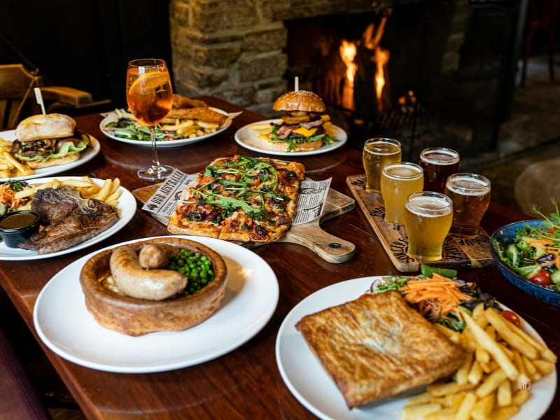 Drewniany stół zastawiony różnorodnymi daniami i napojami w ciepłym świetle kominka.