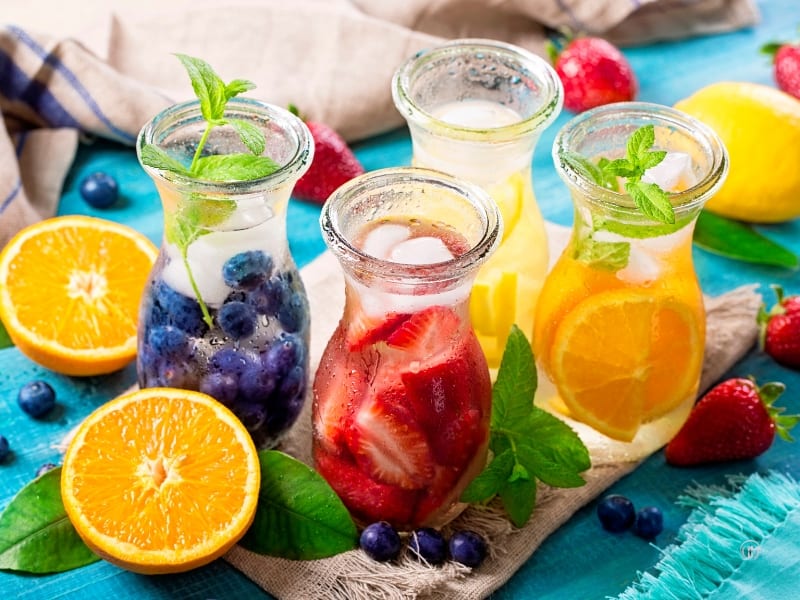 Karafki z napojami owocowymi z cytrusami, truskawkami i jagodami na błękitnym tle.