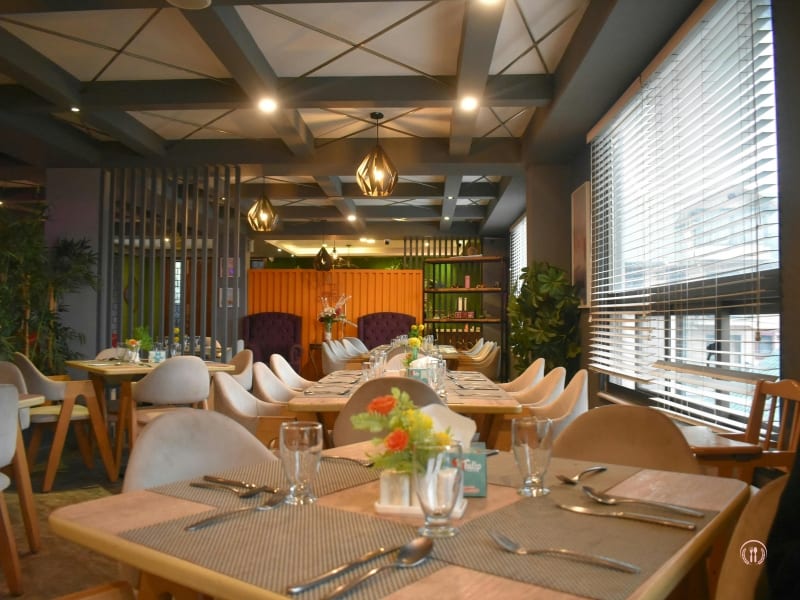 Sala restauracyjna z jasnym drewnem, dużymi oknami i ciepłym światłem nad stołami.