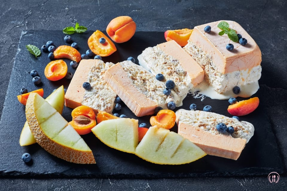 Plasterki semifreddo z mascarpone i owocami leśnymi na ciemnym łupku, z cząstkami melona, pomarańczowymi owocami i borówkami.