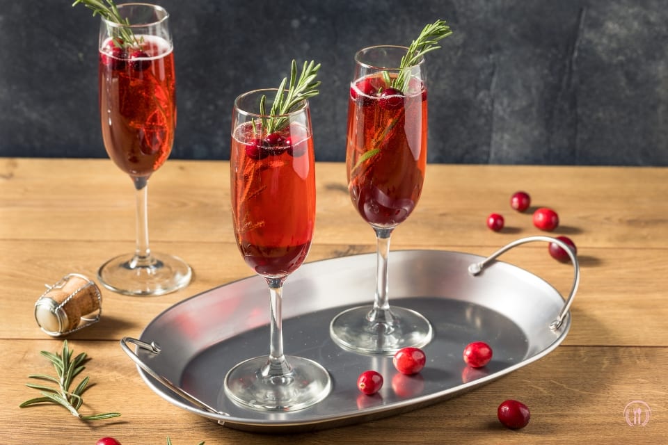 Trzy kieliszki z czerwonym koktajlem szampańskim, ozdobione rozmarynem i żurawiną na metalowej tacy.