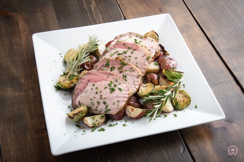 Plastry polędwicy wołowej na białym kwadratowym talerzu, z pieczonymi warzywami i ziołami.
